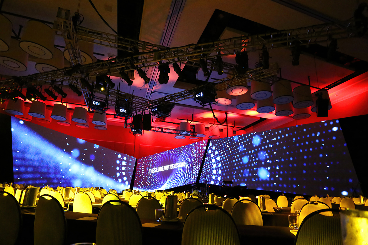Ballroom set up with tables, chairs, a stage, multiple screens, and lighting