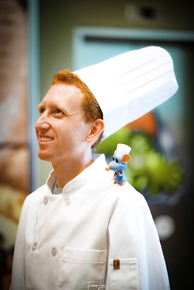 Man in a chef's costume with a toy rat on his shoulder wearing a chef's hat